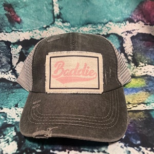 May include: A black and gray distressed trucker hat with a white patch that says "Baddie" in pink lettering.