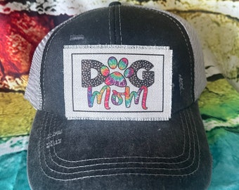 May include: Black and gray distressed trucker hat with a white patch that says "Dog Mom" in rainbow letters with a rainbow paw print in the middle of the word "Dog".