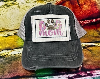 May include: A distressed black baseball cap with a white mesh back. The front features a rectangular patch with the words "Dog Mom" and a paw print design in pink, purple, and brown.