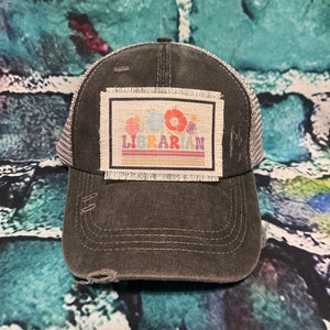 May include: A black and gray distressed trucker hat with a patch that says "Bookworm Librarian" with colorful flowers and a rainbow stripe.
