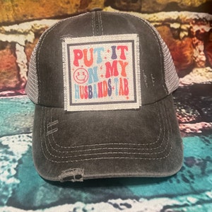 May include: A black and gray distressed trucker hat with a patch that says "PUT IT ON MY HUSBANDS TAB" in colorful letters.