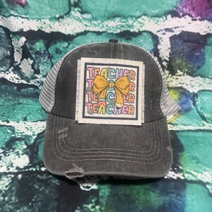 May include: A black and gray distressed trucker hat with a colorful embroidered patch that says "Teacher" with a yellow bow.
