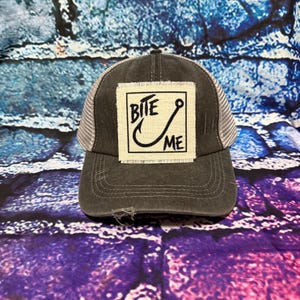 May include: A charcoal gray trucker hat featuring a rectangular off-white fabric patch with frayed edges. The patch displays the black text BITE ME alongside a graphic of a fishing hook. The hat has a mesh back and a distressed brim.
