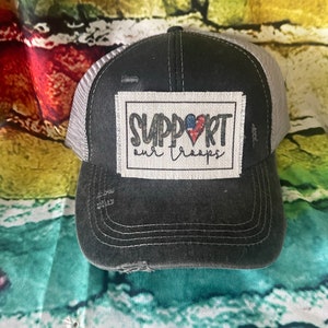 May include: A black and gray distressed trucker hat with a white patch that says "SUPPORT our troops" with a red, white, and blue heart.