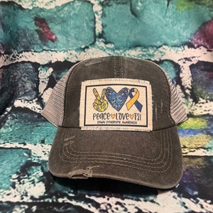 May include: A black and gray trucker hat with a white patch that says "Peace Love T21 Down Syndrome Awareness" and features a peace sign, a heart with a leopard print, and a yellow ribbon.
