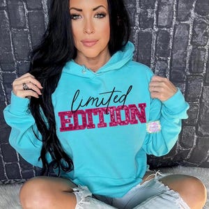 Puede incluir: Una sudadera con capucha azul turquesa con una inscripción cursiva negra "limited" y una inscripción brillante rosa "EDITION".