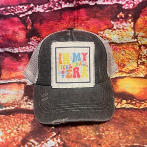 May include: A black and gray mesh trucker hat with a distressed look. The hat has a patch on the front that says "In My Pickleball Era" in colorful letters.