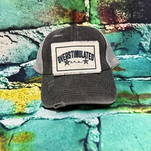 May include: A distressed charcoal gray trucker hat with a white mesh back. The front features a rectangular patch with the words "OVERSTIMULATED era" in black lettering. Two small star graphics flank the word "era".