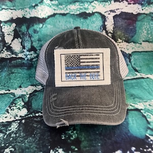May include: A black and gray distressed trucker hat with a white patch featuring an American flag and a blue line. The patch also has the text "Back the Blue" in blue.