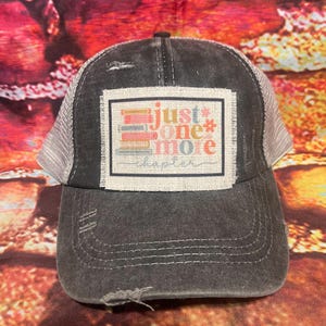 May include: A black and gray trucker hat with a distressed look. The hat has a patch on the front that says "Just one more chapter" with a stack of books in the background.