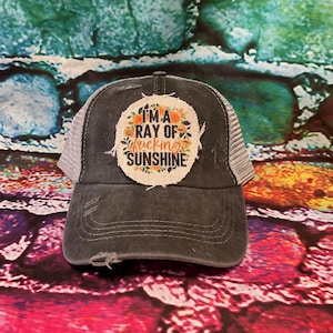 May include: A dark gray trucker hat with a distressed look. The hat has a white patch with the text "I'm a ray of f*cking sunshine" and floral accents.
