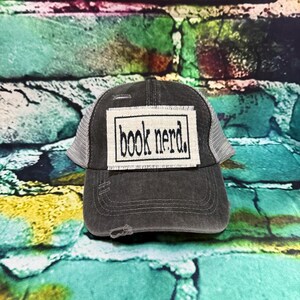 May include: A distressed charcoal gray trucker hat with a white mesh back. The front patch reads "book nerd" in black block letters inside a white rectangle. The hat has a pre-worn look with distressed details.