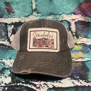 May include: A black and gray trucker hat with a distressed look. The hat has a patch on the front that says "Somebody's Problem" in white lettering with a red background.