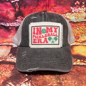 May include: A black and gray distressed trucker hat with a white patch that says "In My Pickleball Era" with a green pickleball and two green paddles.