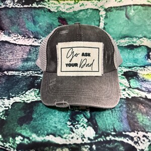 May include: A distressed gray trucker hat with a white mesh back. The front patch reads "Go Ask Your Dad" in black script. The hat has a pre-worn look with a slightly frayed brim, perfect for casual wear.