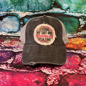 May include: A black and gray trucker hat with a distressed look. The hat has a patch on the front that says "Losing my shit one meltdown at a time". The patch has a floral design around the edges.
