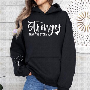May include: Black hoodie with the text "stronger than the storm" in white script. A white heart graphic is next to the word "stronger". A white heart and "P" graphic is on the left sleeve. The hoodie has a front pocket and a hood.