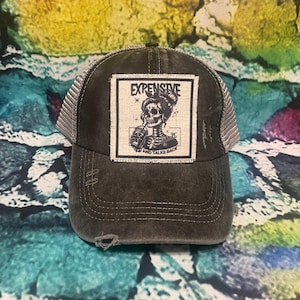May include: A black and gray distressed trucker hat with a white patch featuring a skull with a drink and the text "Expensive and Talks Back".