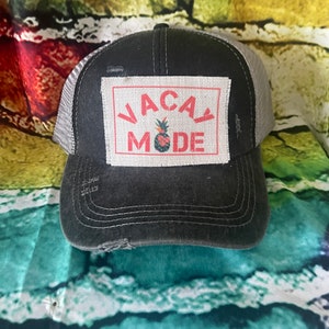 May include: A black and gray distressed trucker hat with a white patch that says "VACAY MODE" in red letters with a pineapple graphic in the middle.