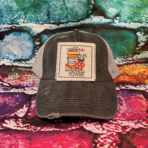 May include: A distressed charcoal gray trucker hat with a white mesh back. The front features a square patch with the text "coffee gives teacher powers" above a stack of books, a coffee cup, and a red apple.