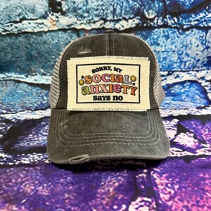 May include: A distressed gray trucker hat with a mesh back and a patch that reads "Sorry, my social anxiety says no" in colorful text. The hat has a curved brim and a worn appearance, suggesting a vintage or casual style.