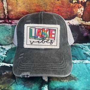 May include: A black and gray distressed trucker hat with a white patch that says "LAKE vibes" in rainbow letters.