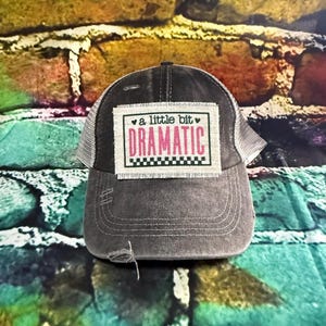 May include: A black and gray trucker hat with a distressed look. The hat has a patch on the front that says "a little bit dramatic" in pink letters with a heart on each side of the text.