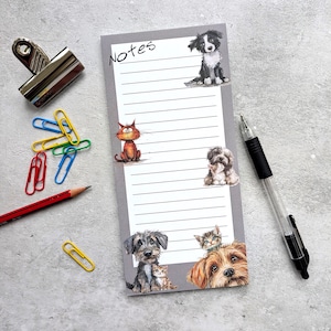 May include: A grey notepad with the word "Notes" at the top, decorated with illustrations of cats and dogs. A silver binder clip, colourful paper clips, a red pencil, and a pen are also visible.