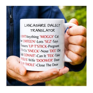 May include: A white ceramic mug featuring the black and red text Lancashire Dialect Translator, listing various regional terms and their definitions, including OWT, MOGGY, UMTEEN, SEZ, UP T'STICK, SNECK, DEE, CONNIT, TEK, MISSUS, DOOWER, and THOLE.