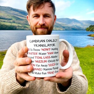 May include: A white ceramic mug featuring the black text CUMBRIAN DIALECT TRANSLATOR at the top, followed by a list of dialect words in red and their English definitions in black, including terms like NOWT, WATTER, YAM, and GINNEL.