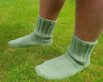 May include: A pair of light green knitted socks with ribbed cuffs. The socks are made of a soft, textured yarn and are folded over at the top. The socks are shown on a grassy background.