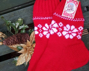 May include: Pair of red knitted Christmas stockings with white snowflake patterns and a white trim. A tag with a Santa Claus illustration hangs from one stocking. The stockings are displayed on a green wooden surface with holiday decorations.