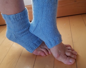 May include: A pair of light blue knitted socks. The socks are a solid color and have a ribbed cuff. The socks are worn on a person's feet, with the feet resting on a wooden floor.