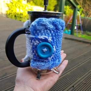 May include: A French press coffee maker with a blue and white knitted cozy. The cozy has a large blue button and is wrapped around the glass carafe. The handle is black and the top is silver.