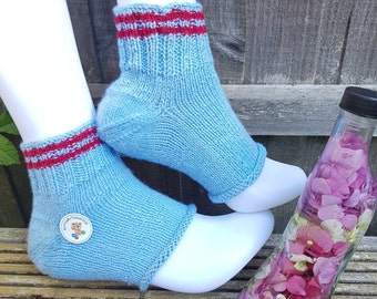 May include: A pair of light blue knitted ankle socks with red stripes at the top. The socks are displayed on white mannequin feet. A small button with a teddy bear design is attached to one sock. A clear bottle filled with pink and purple flowers is next to the socks.
