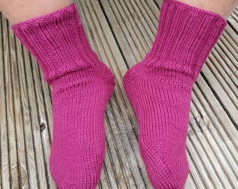 May include: A pair of knitted socks in a vibrant fuchsia color. The socks have a ribbed cuff and a textured knit pattern throughout the leg and foot. The socks are displayed on a wooden surface.
