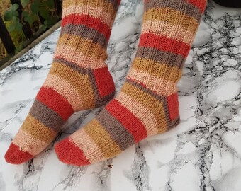 May include: A pair of hand-knitted socks with horizontal stripes in shades of red, brown, tan, and peach. The socks are made of a thick yarn and appear warm and cozy. The socks are resting on a white and gray marble surface.