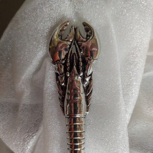 May include: A silver lobster-shaped bottle opener with a detailed design. The opener has a sharp point at the bottom and a realistic lobster claw design at the top.