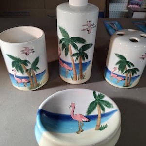 May include: A bathroom set with a pink flamingo theme. The set includes a soap dispenser, toothbrush holder, tumbler, and soap dish. Each piece features a pink flamingo and palm trees on a white background.