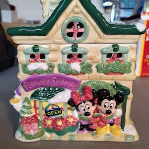 May include: A ceramic cookie jar shaped like a house, featuring a green roof and windows with red accents. The jar is decorated with Minnie and Mickey Mouse characters and the text "Flower Shoppe".