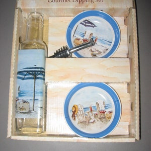 May include: Gourmet dipping set with a clear glass bottle, a black and silver bottle opener, and two blue and white ceramic dishes with beach scenes. The dishes feature beach chairs, an umbrella, and a beach.