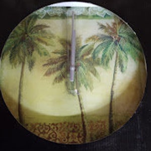 May include: A round, decorative plate with a tropical theme. The plate features a painted design of three palm trees with green leaves and brown trunks against a yellow and green background.