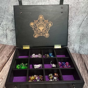 May include: Black wooden dice box with a laser-etched design on the lid. The interior has purple felt lining and compartments filled with various dice. The box has a handle and a latch.