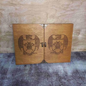 May include: A wooden box with two hinged doors, each featuring a laser-engraved design of a dragon with a shield and axes. The box has metal hinges and clasps.