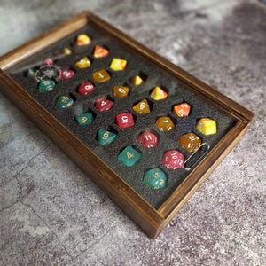 May include: A wooden dice tray containing a collection of multi-sided dice in various colours, including green, red, yellow, and orange. The dice have white or gold numerals. A silver safety pin is also present.