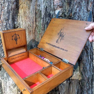 May include: A wooden gaming box with a hinged lid, featuring a red felt-lined interior with multiple compartments for dice. The lid displays a black graphic of a figure and the text, "He will be laughing still... at the end."