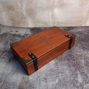 Peut inclure: Une boîte rectangulaire en bois avec une finition brun rougeâtre. La boîte présente des coins de protection en laiton, des sangles en cuir noir et un emblème ovale avec un motif orné sur le couvercle. La boîte est fermée.