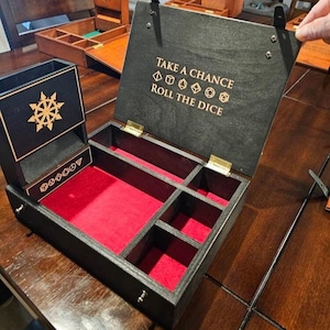 May include: A black wooden dice box with a red velvet interior, featuring a built-in dice tower and multiple compartments. The lid interior is engraved with the text "TAKE A CHANCE ROLL THE DICE" and icons of various polyhedral dice shapes.