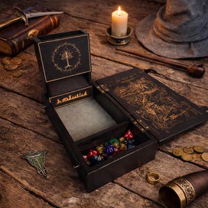 May include: A black wooden dice box with gold accents, featuring a tree and text design. The box is open, revealing colorful dice. Other items include a candle, a hat, a pipe, and a book with gold illustrations.