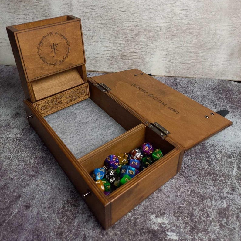 Op de afbeelding: Houten dobbelbox met meerdere compartimenten. De doos is open en toont een verzameling kleurrijke dobbelstenen. Op het deksel staat de tekst "please let me win". Het bovenste gedeelte heeft een gegraveerd ontwerp en een symbool.